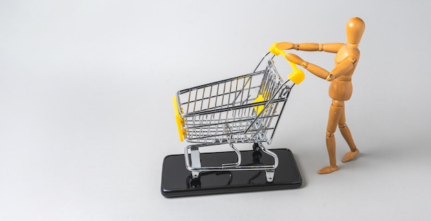 Деревянная человеческая фигура с миниатюрной тележкой для покупок на мобильном телефоне Концепция онлайн-покупок