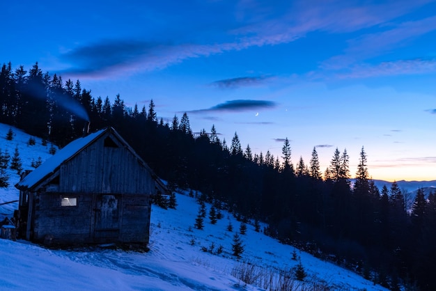 A wooden hut standing in the mountains in winter night landscape smoke from the chimney of the hut