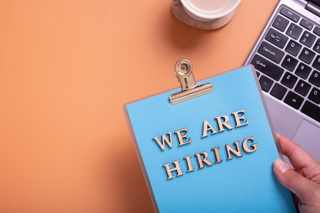 Wooden letters with phrase we are hiring on clipboard in female hand on color background with laptop and coffee Search worker concept