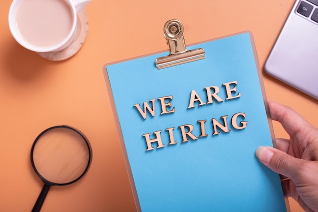 Wooden letters with phrase we are hiring on clipboard in female hand on color background with laptop magnifire and coffee Search worker concept