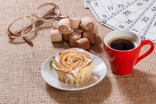 Wooden lotto barrels and game cards for a game in lotto with glasses, cup of coffee and homemade biscuit with apples in the form of rose on white saucer