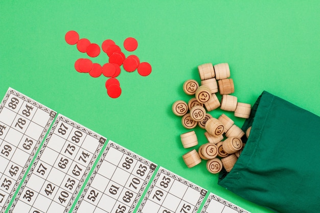 Photo wooden lotto barrels with cloth bag, game cards and red chips on green background. board game lotto. top view with copy space