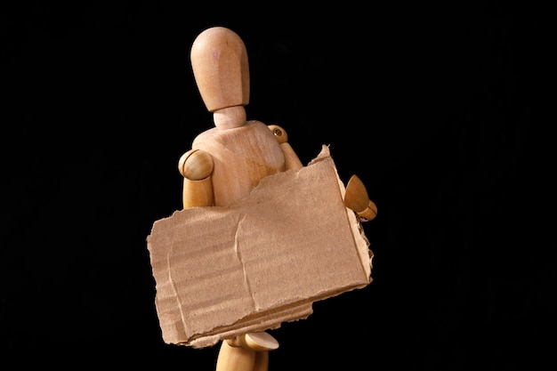 A wooden man holds a brown cardboard sign on a black background High quality photo