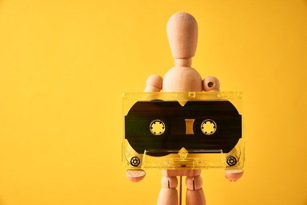 Wooden man holds retro cassette tape on yellow