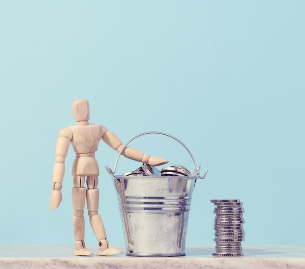 A wooden mannequin and coins in a miniature bucket on a blue background a concept of high income subsidy