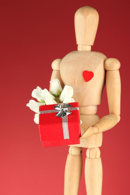 Wooden mannequin holding gift box and bouquet on red background