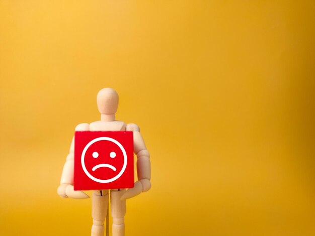 Wooden mannequin holding red cube with sad face on a yellow background with copy space