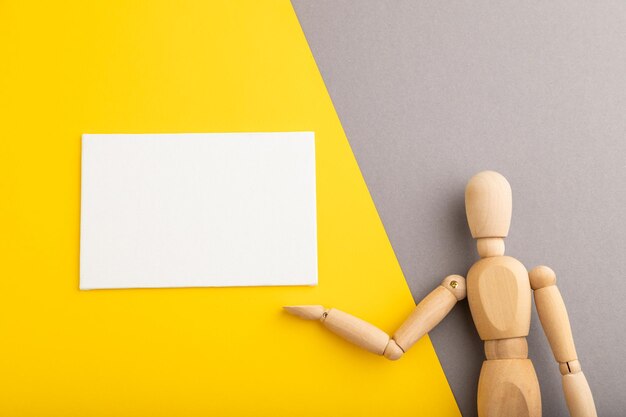 Wooden mannequin holding white blank poster on gray and yellow pastel background copy space