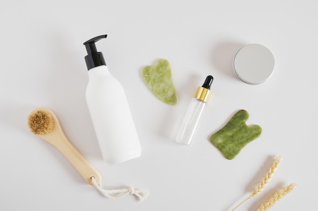 Wooden massage brush, aluminum cosmetics jar, cream in a bottle\
with a dispenser, serum in dropper bottle and a gua sha scraper\
made of jadeite stone