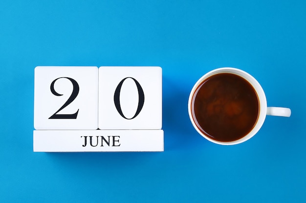 Деревянный блокнот с датой 20 июня и кружкой кофе на голубом пастельном фоне.