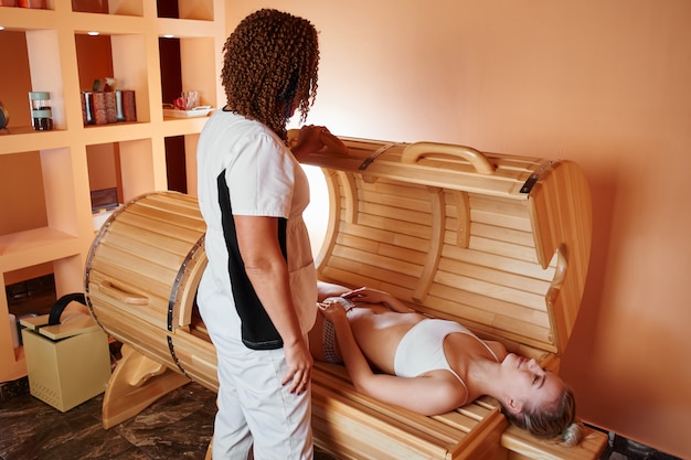 Photo wooden phyto barrel, horizontal sauna. beautiful woman massage therapist applying spa treatment to a young woman in a wellness room