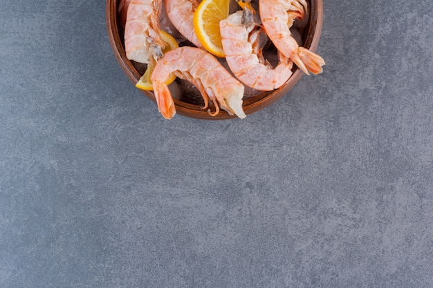 A wooden plate of delicious shrimps with ice cubes and sliced lemon on a stone surface