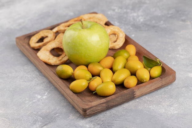 Wooden plate of fresh cumquats, apple and dried apple rings on marble.