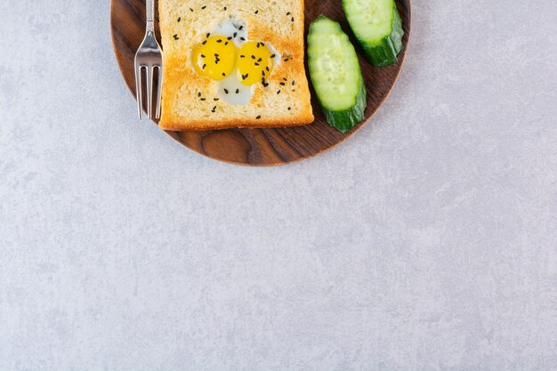 Wooden plate of toast with fried eggs and cucumbers on table.