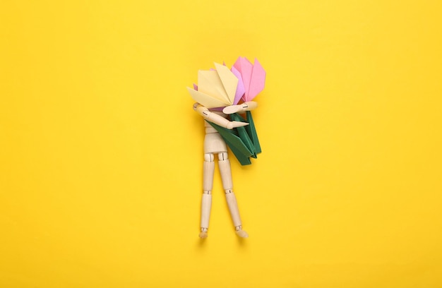 Wooden puppet holds origami tulips on a yellow background Top view