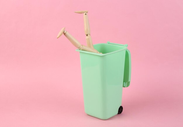 Wooden puppet rummages in a trash can on a pink background Poverty concept