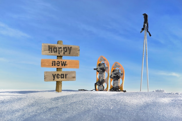 Cartello in legno con testo felice anno nuovo sulla neve accanto a racchette da neve e bastoncini da sci