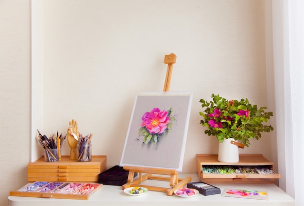 wooden sketch easel of a pink rosehip flower stands on a table next to the materials for drawing with brushes, pencils and dry pastel. concept of the workplace of a female artist. copyspace