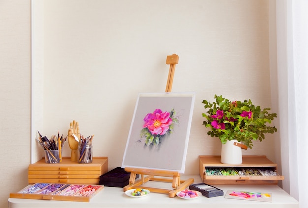 Wooden sketch easel of a pink rosehip flower stands on a table next to the materials for drawing with brushes, pencils and dry pastel. Concept of the workplace of a female artist. Copyspace