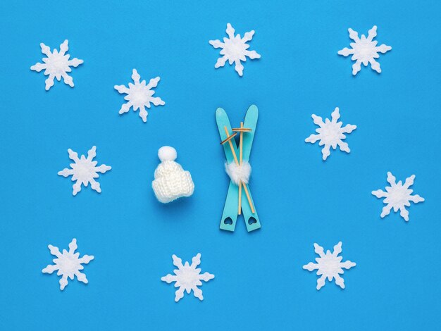 Photo wooden skis with sticks and a knitted hat on a blue background with snowflakes. flat lay.