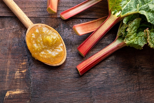 A wooden spoon with homemade rhubarb jam and fresh oraganic rhubarb stalks on a dark old table