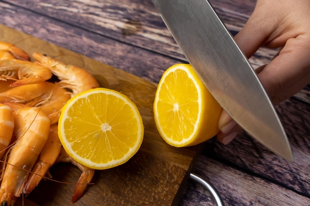 On a wooden stand a lemon is cut with a knife near boiled shrimp