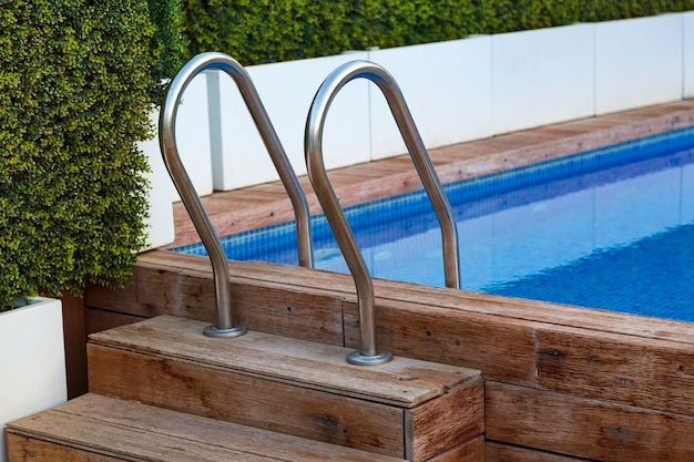 Wooden steps into the pool with metal handrails The planking of a swimming pool with stair and turquoise clear water Open air swimming pool with wooden deck stainless stair and blue surface