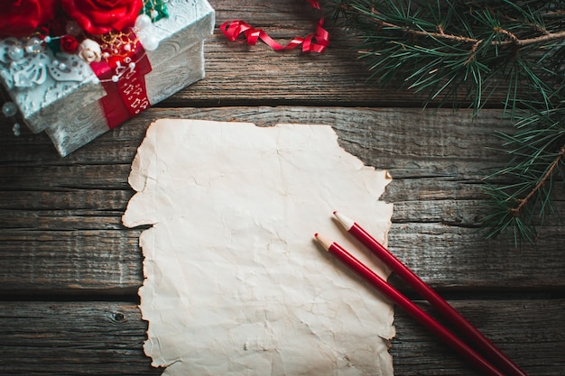 На деревянном столе - старая желтая бумага, красная, карандаши, подарок и ветки деревьев на новый год.