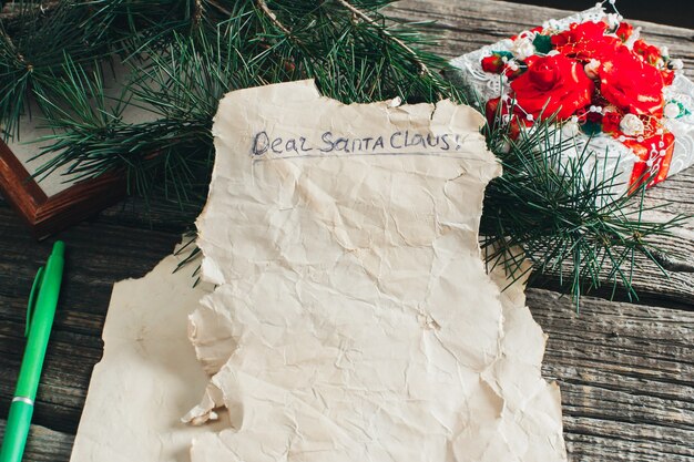 On the wooden table are an old yellow paper that says, Dear Santa Claus red, pencils, pens assorted colors gift and tree branches for the new year