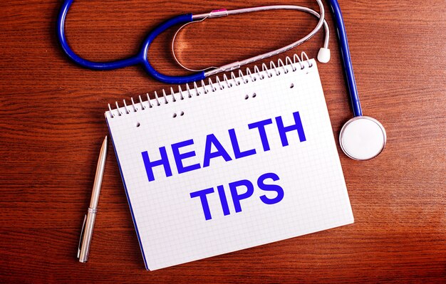 On a wooden table are a pen, a stethoscope, and a notebook labeled HEALTH TIPS. Medical concept.