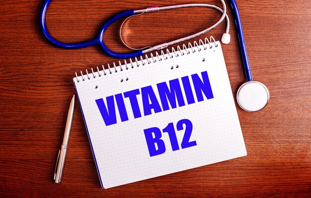 나무 테이블에는 펜, 청진기, VITAMIN B12라고 표시된 노트북이 있습니다.