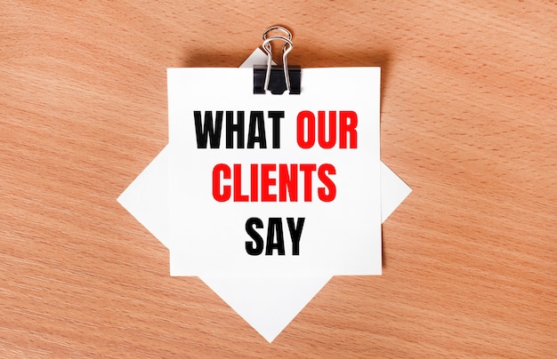 On a wooden table under a black paper clip lies a sheet of white paper with the text WHAT OUR CLIENTS SAY