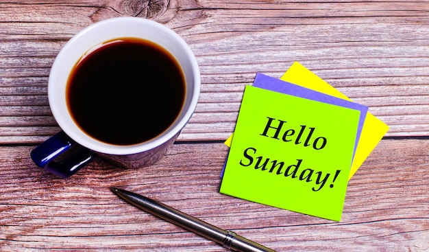 Photo on a wooden table - a blue cup with coffee, a pen and colorful bright stickers with the text hello sunday. view from above