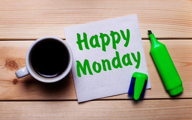 On a wooden table, a cup of coffee, a green marker and a napkin with the text HAPPY MONDAY