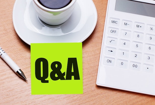 The wooden table has a white cup of coffee, a pen, a white\
calculator, and a green sticker with the text q and a. business\
concept