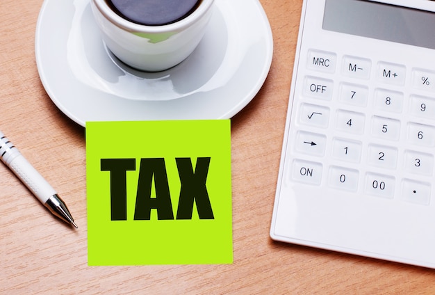 The wooden table has a white cup of coffee, a pen, a white calculator, and a green sticker with the text TAX. Business concept