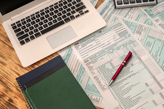 On a wooden table lie a laptop, American tax forms with a tablet and a pen. Documentation concept