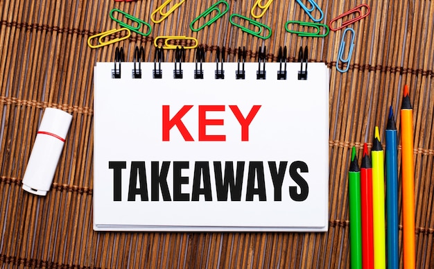 Su un tavolo di legno, matite multicolori, graffette, una chiavetta usb e un quaderno con il testo key takeaways. lay piatto