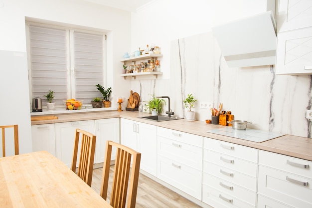 Wooden table in sunny and bright kitchen with white marble wall. Scandinavian design kitchen with plants, accessories and straw bag.