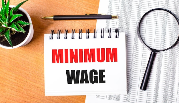 나무 테이블에는 보고서, 화분, 돋보기, 검은색 펜 및 최소 임금(MINIMUM WAGE)이라는 텍스트가 있는 공책이 있습니다. 비즈니스 개념