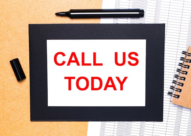 On a wooden table, there is a black open marker, a brown notepad and a sheet of paper in a black frame with the text CALL US TODAY. View from above.