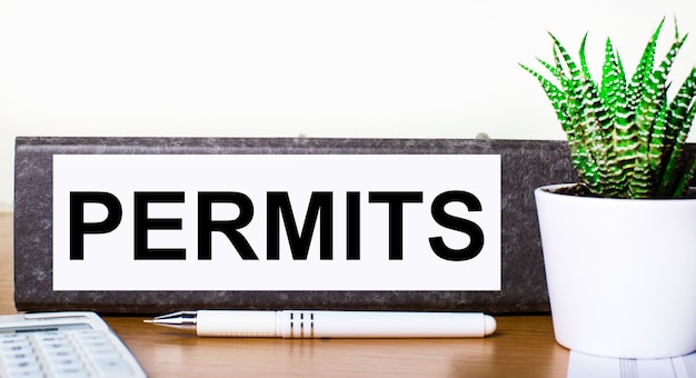 On a wooden table there is a folder for documents with the text PERMITS, a green plant in a pot, a pen and a calculator. Business concept.