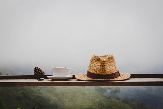 On a wooden table, there is a hot coffee cup and a hat placed in the wilderness.