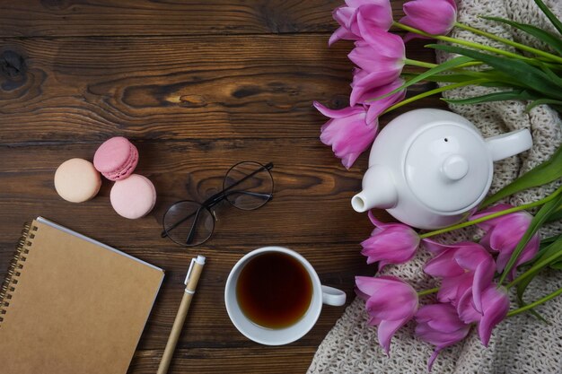 On a wooden table there is a white teapot and a cup of tea a notebook and a bouquet of tulips