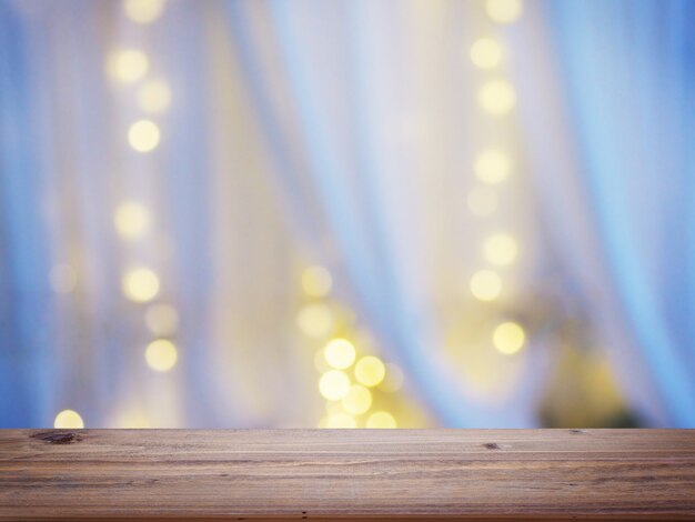 Wooden table top over abstract blur background of white curtain with light bulb bokeh at window in bedroom.