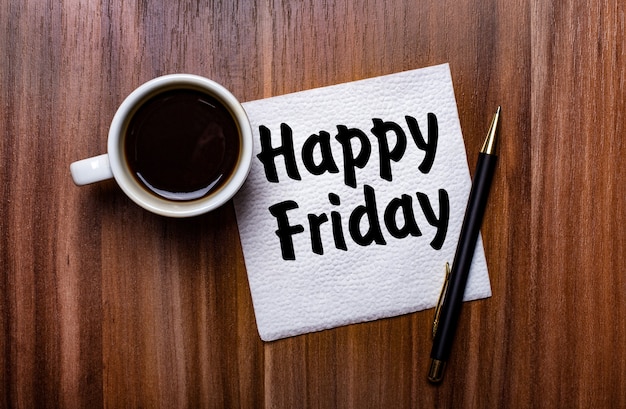 Photo on a wooden table next to a white cup of coffee and a pen is a white paper napkin with the words happy friday