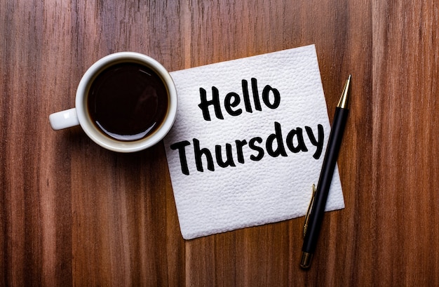 On a wooden table next to a white cup of coffee and a pen is a white paper napkin with the words HELLO THURSDAY