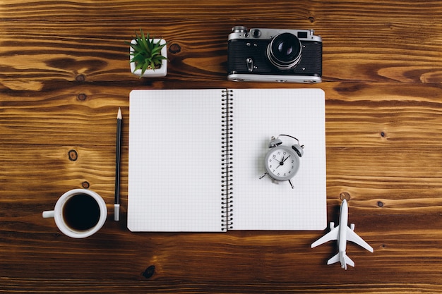 Wooden table with cup of coffee, alarm clock, airplane, camera, notebook and green. 