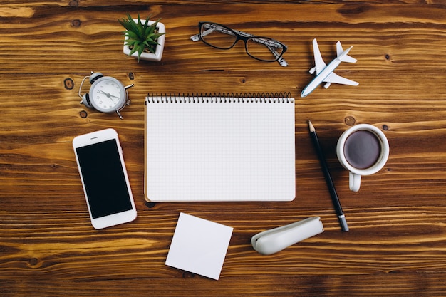 Wooden table with cup of coffee, alarm clock, smartphone, airplane, glasses, notebook