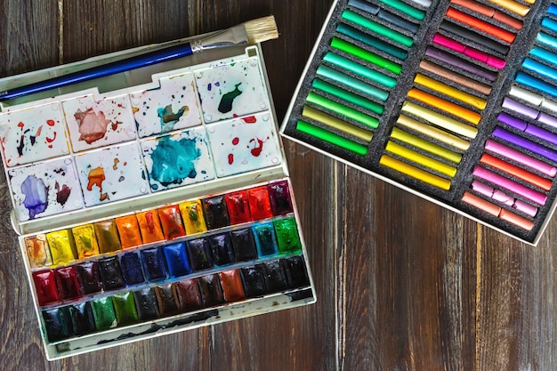 wooden table with paint brushes, watercolor paints and crayon pastel chalk